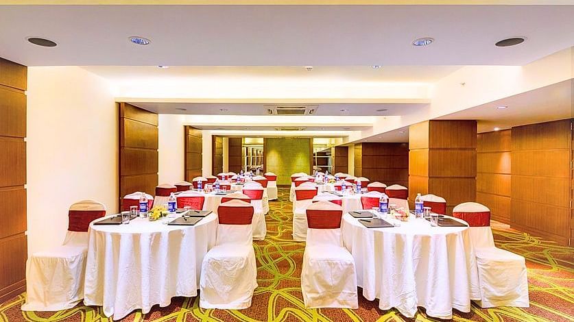 A spacious banquet hall at Pride Plaza Ahmedabad, featuring multiple tables with white tablecloths and red chair covers, set for a formal event or conference.